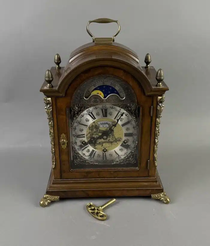 Kommodenuhr John Warmink im Barockstil Zifferblatt mit Mondphasenanzeige, römische Ziffern, braun mattiertes Holzgehäuse mit Gelbmetallapplikationen, Höhe ca. 39 cm, 1 Schlüssel,